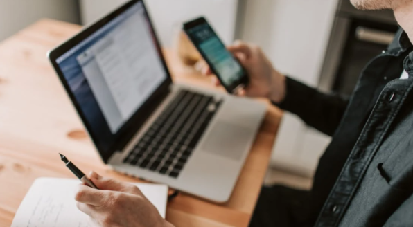 homem com jaqueta preta segura uma caneta e um celular em cada mão. uma mão apoia um caderno, e ele fica em frente a um notebook